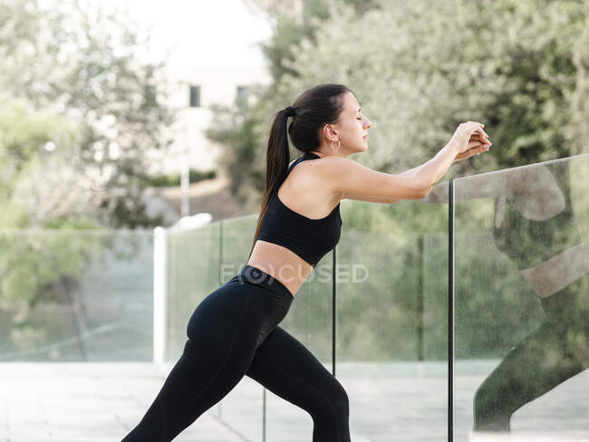 Вид збоку на жіночий бігун у спортивному одязі, що спирається на скляний паркан та легені під час тренувань з фітнесу в літній день — стокове фото