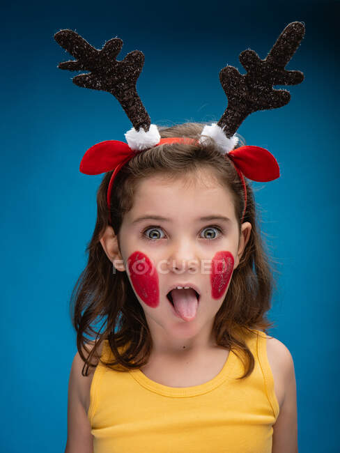 Überraschtes Mädchen mit rot geschminkten Wangen mit Spielzeug-Hirschhörnern und -Ohren und Blick in die Kamera auf blauem Hintergrund, während sie ihre Zunge herausstreckt — Stockfoto