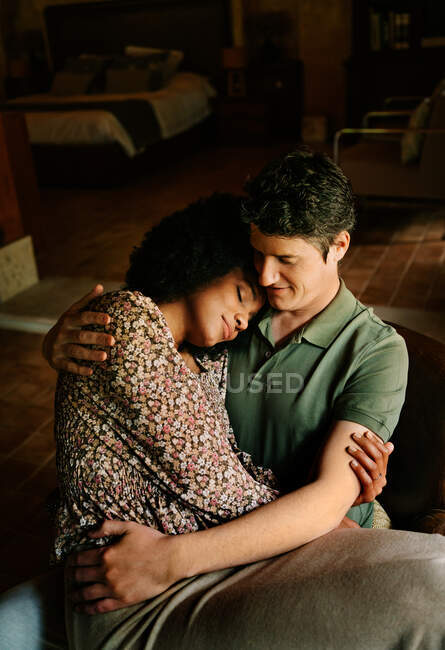 From above of loving black woman with eyes closed sitting on knees of man while embracing at home — Stock Photo