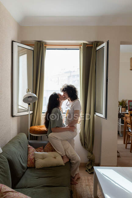 Вид сбоку на содержание ЛГБТ пара женщин целуются на диване дома и смотрят друг на друга с любовью — стоковое фото