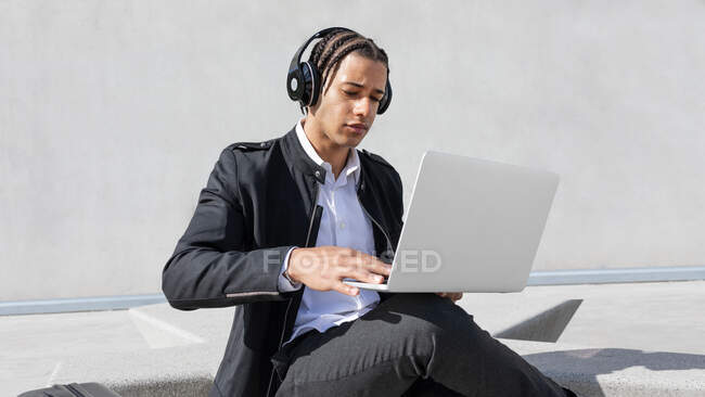 Jovem focado gerente masculino afro-americano com tranças em terno formal e fones de ouvido sem fio enquanto trabalhava em laptop na rua urbana — Fotografia de Stock
