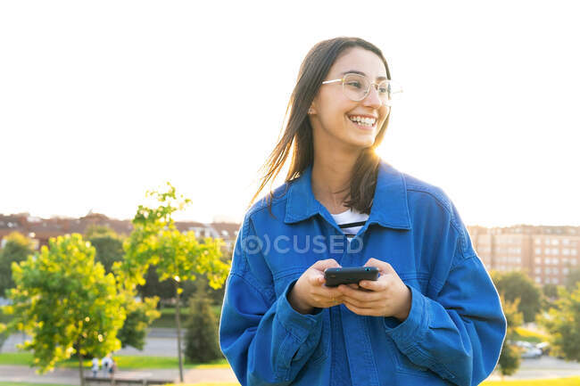 Stilvolle Frau steht auf grünem Grashügel und surft an sonnigen Tagen auf dem Smartphone — Stockfoto