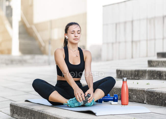 Jovem atleta feminina de sportswear preto ouvindo música em fones de ouvido sem fio verdadeiros e fazendo exercício de aquecimento enquanto se senta em passos na rua da cidade — Fotografia de Stock
