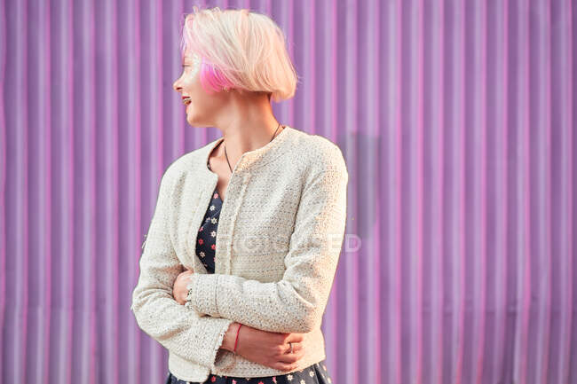 Mujer alternativa encantadora con el pelo teñido y en ropa de moda de pie contra la pared violeta en la ciudad y mirando hacia otro lado - foto de stock