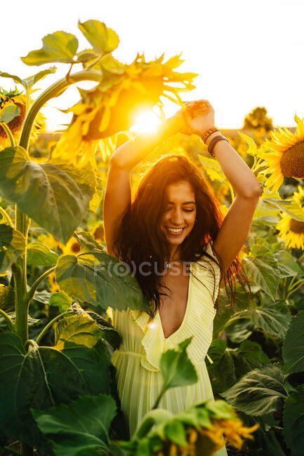 Anmutige, glückliche junge hispanische Frau in stylischem gelben Kleid steht mit erhobenen Armen inmitten blühender Sonnenblumen auf einem Feld in sonnigem Sommertag mit geschlossenen Augen — Stockfoto