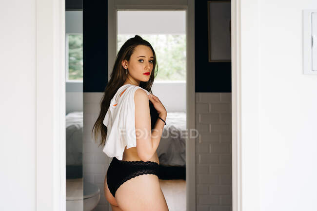 Вид сбоку соблазнительной женщины в черном нижнем белье, стоящей в дверях спальни дома — стоковое фото