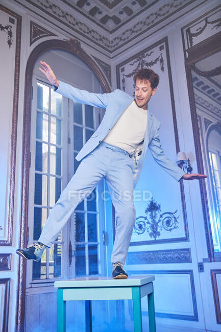 Снизу выразительный счастливый мужчина в стильном костюме балансирует на столе в шикарной комнате с голубым неоновым светом, смотрящим в сторону — стоковое фото