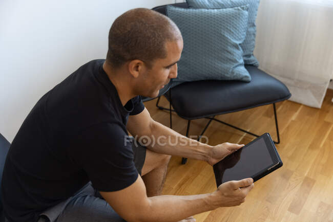 Side view of focused adult male sitting in armchair near table with glass of orange juice and using tablet in morning at home — Stock Photo
