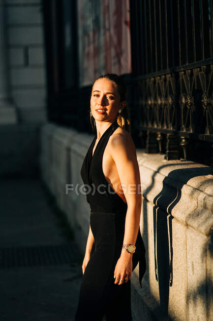 Vista lateral da mulher feliz vestindo roupas da moda em pé na cidade no dia ensolarado e olhando para a câmera — Fotografia de Stock