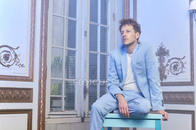 Pensativo hombre elegante en traje de mesa de estar en la habitación elegante y mirando hacia otro lado - foto de stock