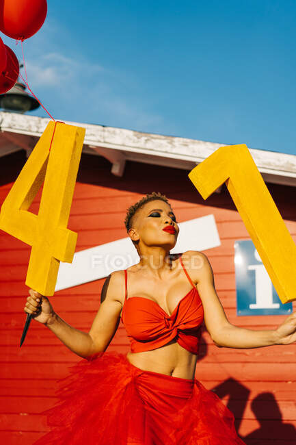 Trendy black female with decorative numbers and red balloons standing on walkway against construction during birthday party in sunlight — Stock Photo