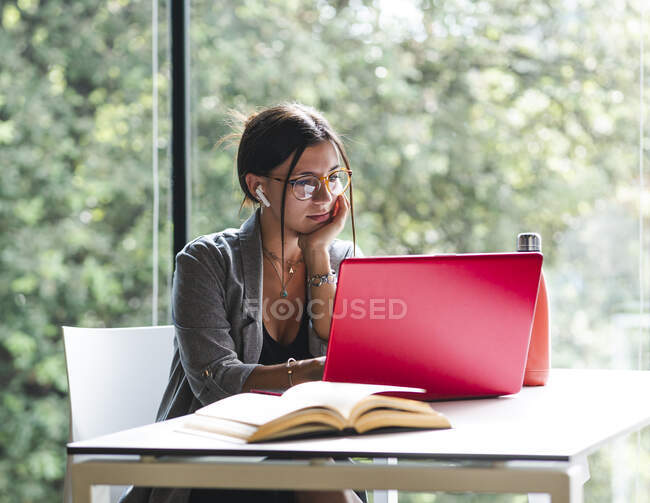 Side view of female student typing on netbook while sitting at table with book and doing homework in university library — Stock Photo
