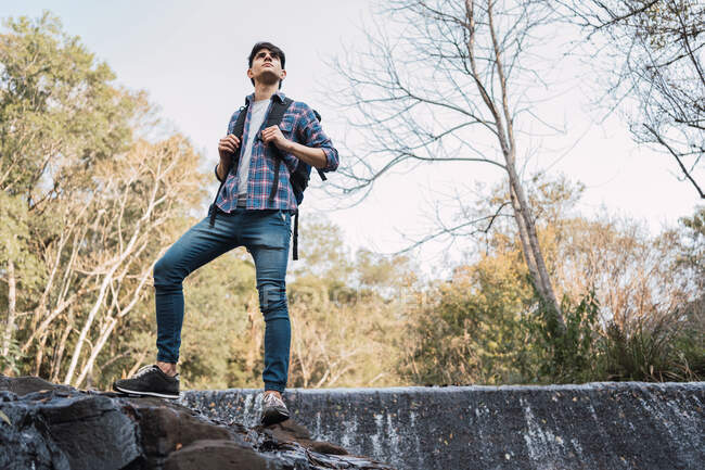 Мужчина турист с рюкзаком стоит на скалистой земле возле водопада в лесу и смотрит в сторону — стоковое фото