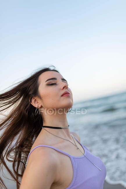 Jovem morena com olhos fechados tremendo cabelo longo contra o mar e céu sem nuvens pôr do sol — Fotografia de Stock