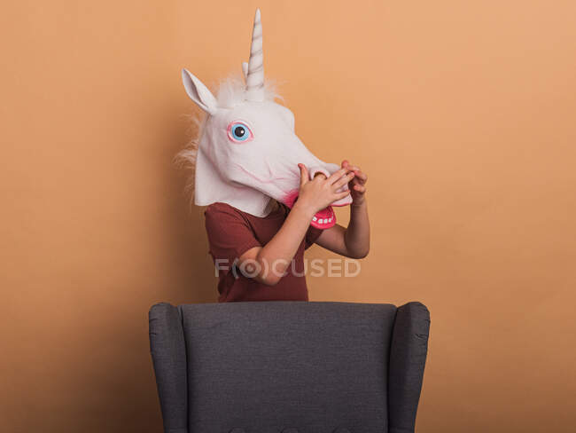 Criança anônima em máscara decorativa unicórnio com boca aberta no fundo bege ao tocar o nariz — Fotografia de Stock