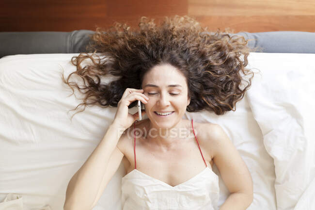 Вид зверху на ділову жінку з кучерявим волоссям, що лежить у ліжку, розмовляючи по телефону — стокове фото