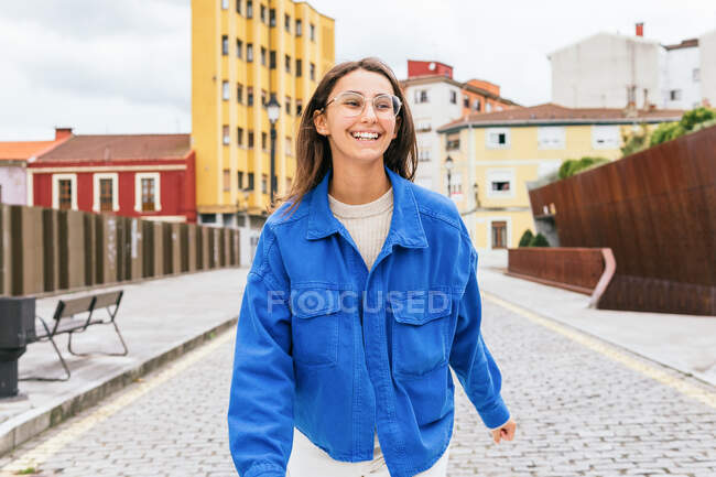 Mujer sonriente con el pelo volador caminando por la carretera en la ciudad en un día ventoso - foto de stock