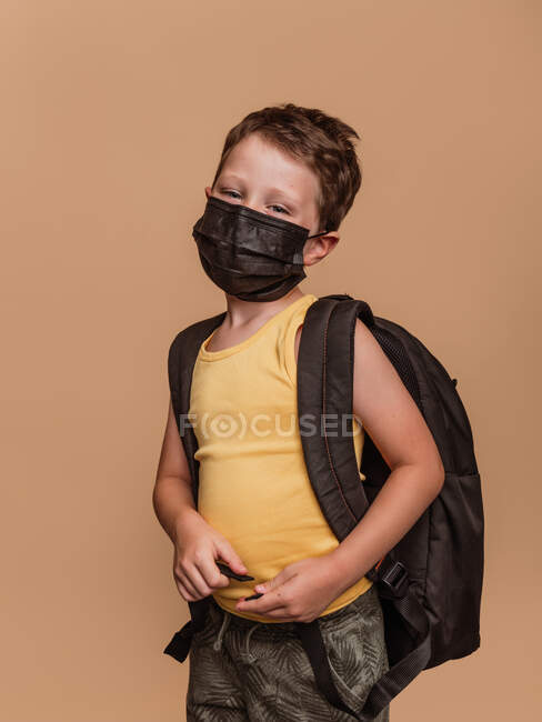 Konzentriertes frühkindliches Schulkind mit Rucksack und in medizinischer Schutzmaske vor Coronavirus blickt in die Kamera auf braunem Hintergrund im Studio — Stockfoto
