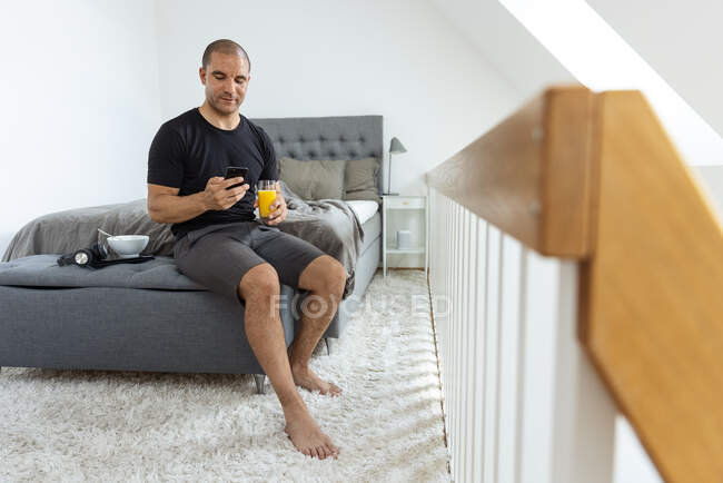 Mann sitzt morgens beim Frühstück zu Hause auf dem Bett und surft mit Handy — Stockfoto