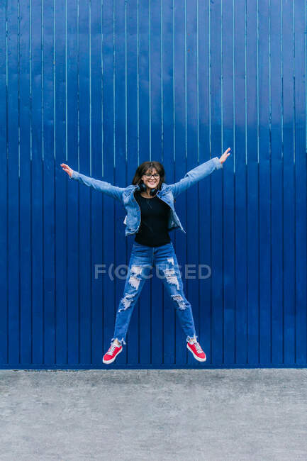 Возбужденная женщина-хипстер в джинсовой одежде прыгает с протянутыми руками на синем фоне в городе и смотрит в камеру — стоковое фото