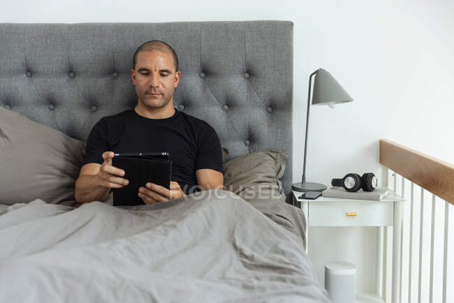 Спокійний чоловік сидить на ліжку під ковдрою і використовує таблетку після пробудження в спальні вранці вдома — стокове фото