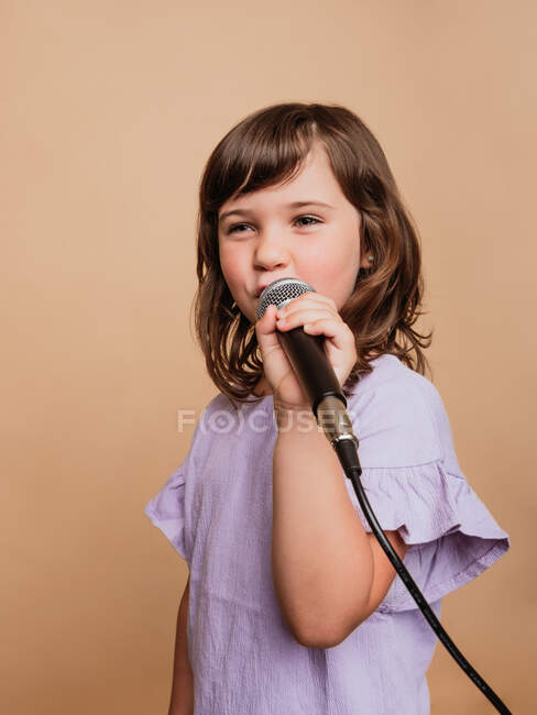 Comic preteen ragazza cantare canzone in microfono su sfondo marrone in studio e guardando altrove — Foto stock