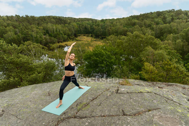 Femme en vêtements de sport noirs levant le bras et faisant Virabhadrasana pose sur un terrain pierreux tout en pratiquant le yoga dans la campagne — Photo de stock
