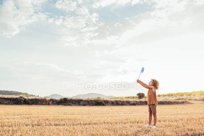 Vista laterale del bambino dai capelli ricci in piedi nel prato asciutto e giocare con il mulino a vento giocattolo in estate — Foto stock
