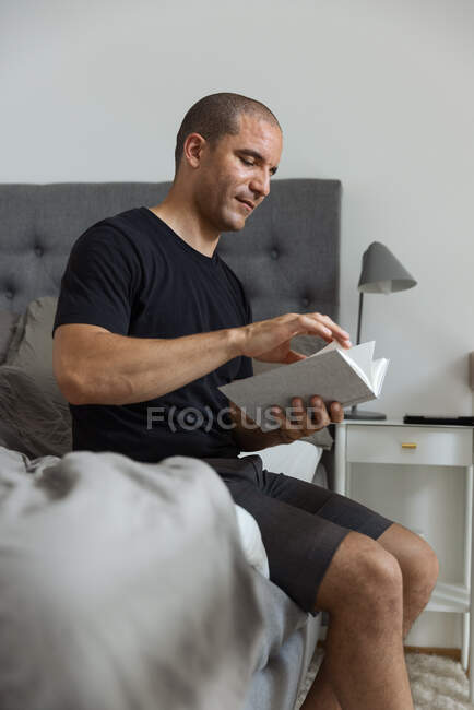 Vista lateral do homem pensativo sentado em cama macia de manhã e lendo história interessante no livro após o despertar — Fotografia de Stock