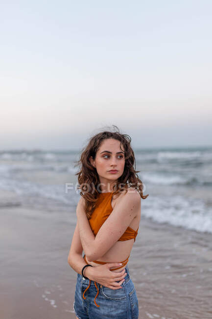 Мрійлива молода жінка з довгим волоссям, дивлячись геть, стоячи на піщаному пляжі біля махаючого моря — стокове фото