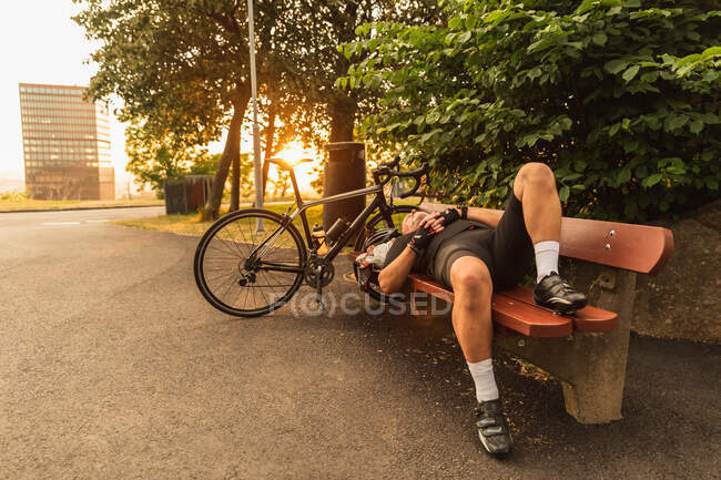 Middle aged ethnic male bicyclist in sportswear lying on city bench against bike and building — Stock Photo
