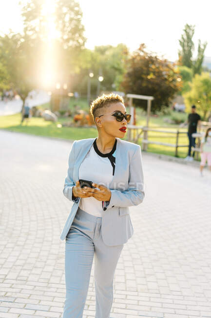 Stylish African American female in sunglasses text messaging on cellphone while strolling on walkway in urban park — Stock Photo