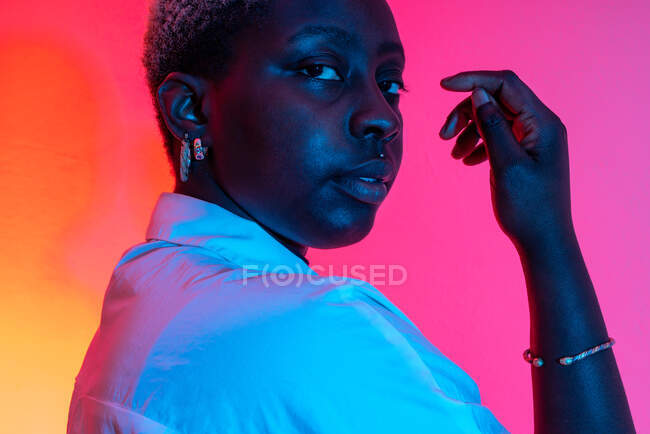 Vue latérale du modèle féminin afro-américain délicat en boucles d'oreilles tendance en studio avec lumière au néon sur fond rose et orange — Photo de stock