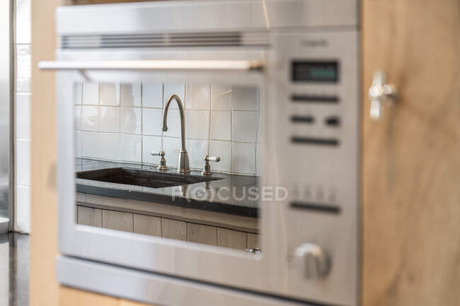 Металлический кран и подмонтированная раковина отражаются в стеклянной двери встроенной духовки в современной кухне в квартире — стоковое фото