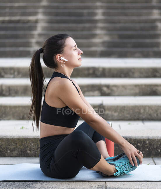Молода жінка - атлет у чорному спортивному вбранні слухає музику у справжніх бездротових навушниках і робить теплі вправи, сидячи на сходах на вулицях міста. — стокове фото