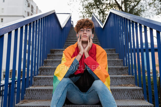 Desde abajo de elegante gay masculino con bandera LGBT sobre hombros sentados en escaleras de metal en la ciudad y mirando a la cámara - foto de stock