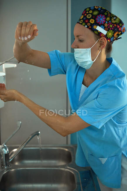 Medico femminile adulto in maschera sterile e uniforme che applica disinfettante liquido a portata di mano sopra il lavandino sul lavoro in ospedale — Foto stock