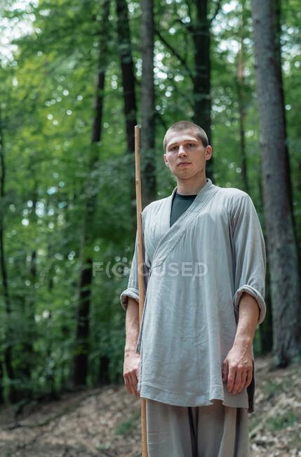 Uomo con bastone che tiene la mano vicino al petto mentre pratica il kung fu nel bosco guardando la fotocamera — Foto stock