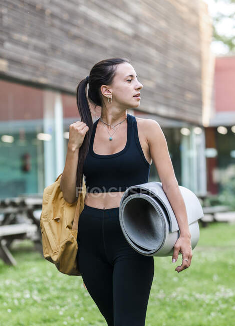 Jeune athlète féminine en tenue de sport noire portant sac et tapis et regardant loin tout en marchant dans la cour herbeuse avant l'entraînement — Photo de stock