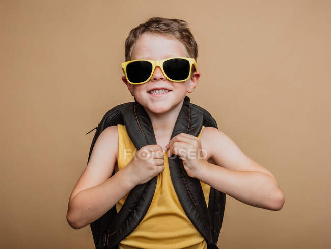 Positif cool écolier pré-adolescente dans des lunettes de soleil et avec sac à dos regardant la caméra sur fond brun en studio — Photo de stock