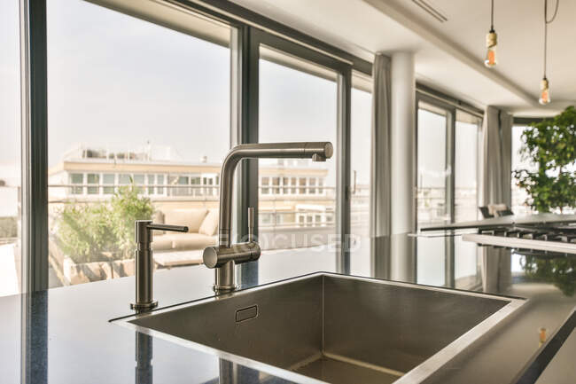 Detalle del diseño interior de la cocina moderna con grifo de cromo y lavabo instalado en un mostrador brillante frente a una gran ventana con vistas al paisaje urbano en un apartamento de estilo loft - foto de stock