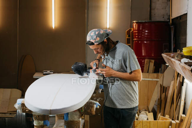Shaper masculino usando plaina elétrica e superfície de polimento de prancha de surf na oficina — Fotografia de Stock
