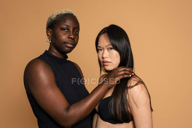 Многорасовые модели женщин в черном нижнем белье с одной рукой на плече другой на бежевом фоне для концепции положительности тела в студии — стоковое фото