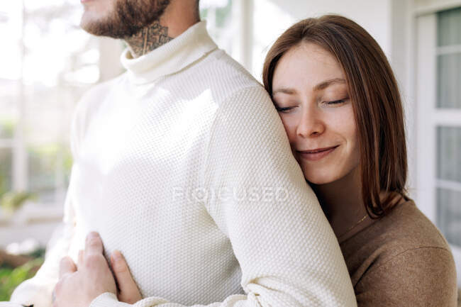 Side view of cheerful female embracing bearded male beloved while looking away against window in house — Stock Photo
