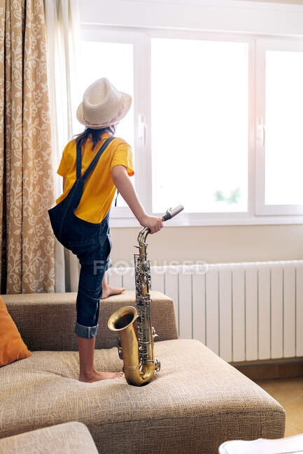Back view of anonymous thoughtful barefoot child in hat with saxophone standing on sofa against window at home — Stock Photo