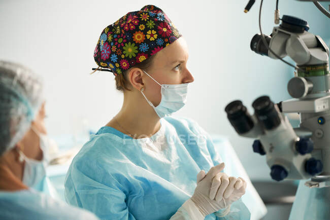 Adult female doctor in surgical uniform and sterile mask looking away while sitting in clinic — Stock Photo