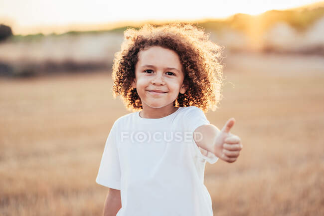 Allegro bambino etnico con i capelli ricci che mostrano come gesto mentre in piedi in campo secco in estate nella parte posteriore illuminata e ammiccando alla fotocamera — Foto stock