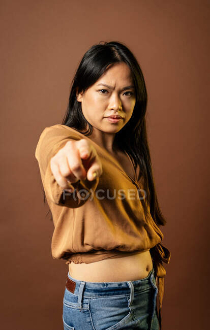 Решительная нахмуренная азиатка, указывающая на камеру и показывающая вам жест на бежевом фоне в студии — стоковое фото