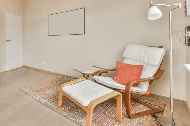 Detalle de diseño interior de apartamento moderno con silla de madera cómoda con cojín suave y reposapiés colocados en la alfombra cerca de la lámpara en la habitación con paredes de luz y cuadro de maqueta - foto de stock