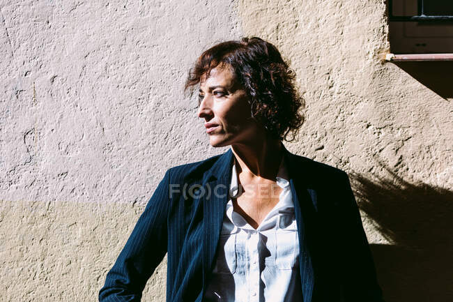 Femme réfléchie portant des vêtements élégants debout la lumière du soleil et jetant l'ombre sur un mur minable et détournant les yeux — Photo de stock